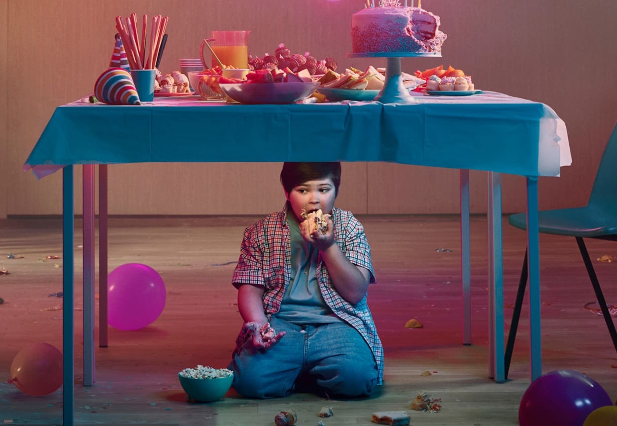 Boy sitting under a table and eating at a birthday party. Actor portrayal.